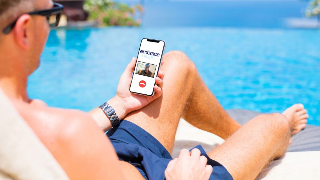 2 A man sits in a chair by a pool holding a phone and talking to a therapist from Embrace Health.